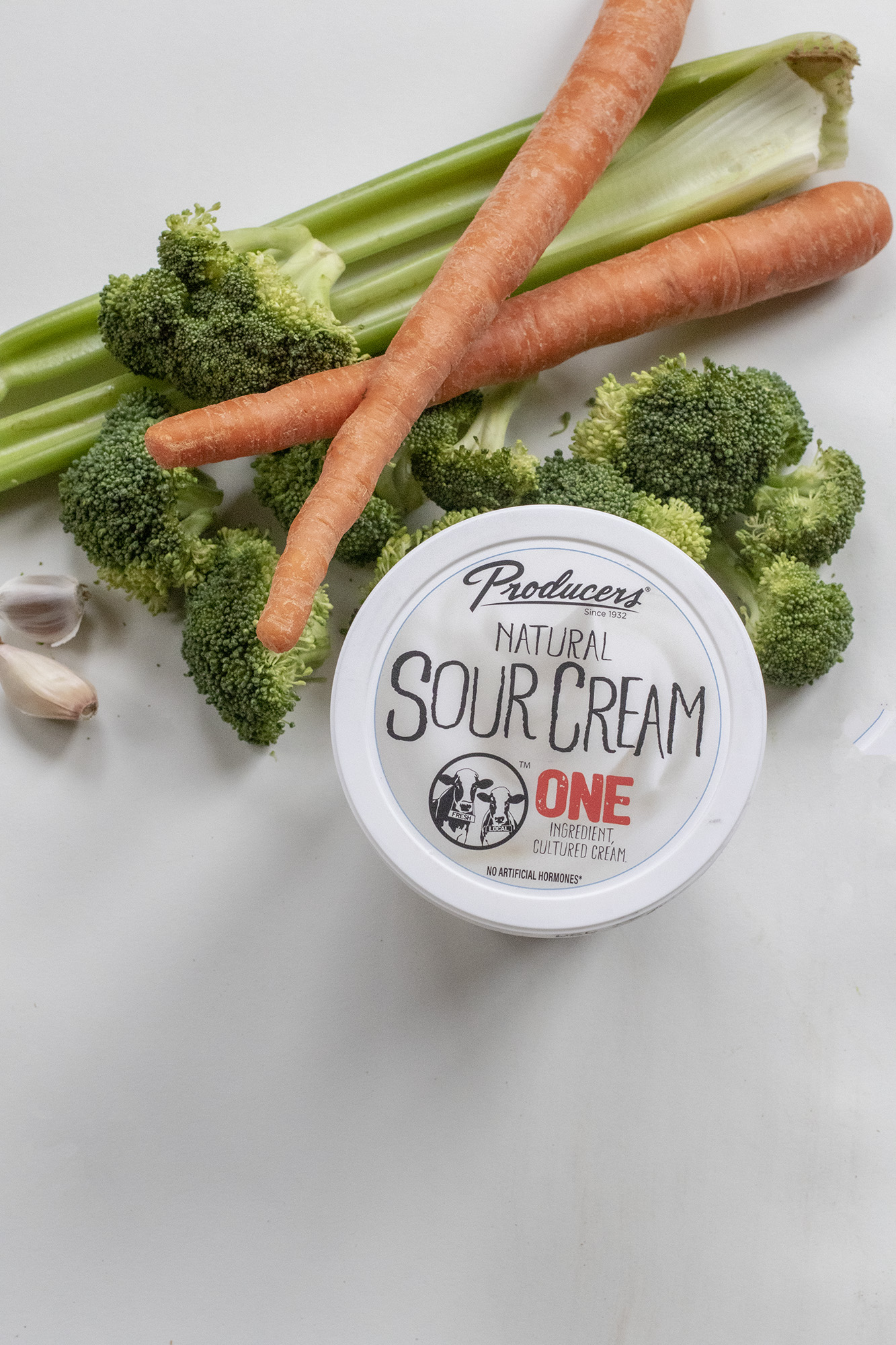 Producers Natural Sour Cream container surrounded by celery, carrots, broccoli and garlic laying flat on a white background.